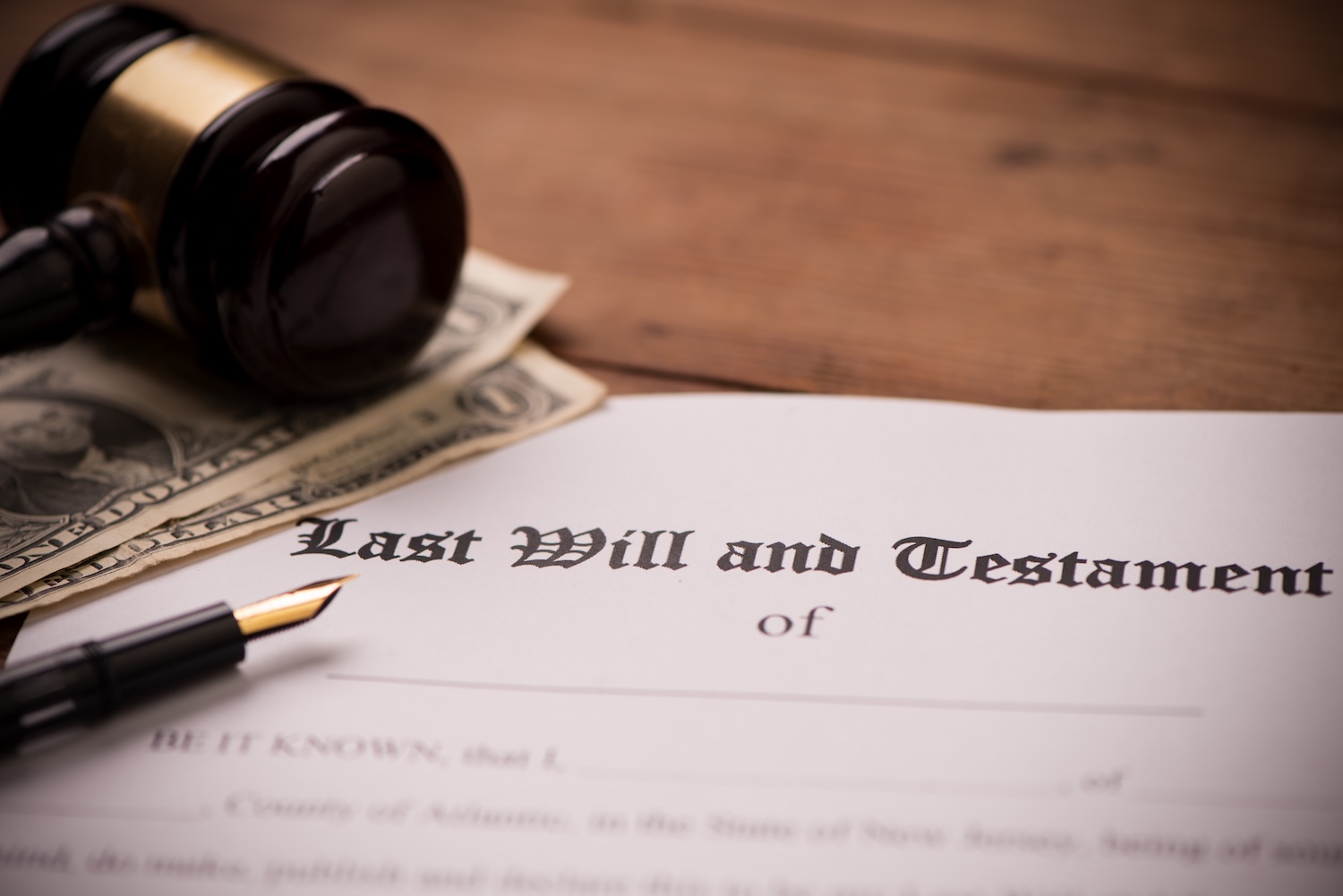 A close-up of a “Last Will and Testament” document on a wooden table with a fountain pen, several one-dollar bills, and a judge’s gavel resting nearby.