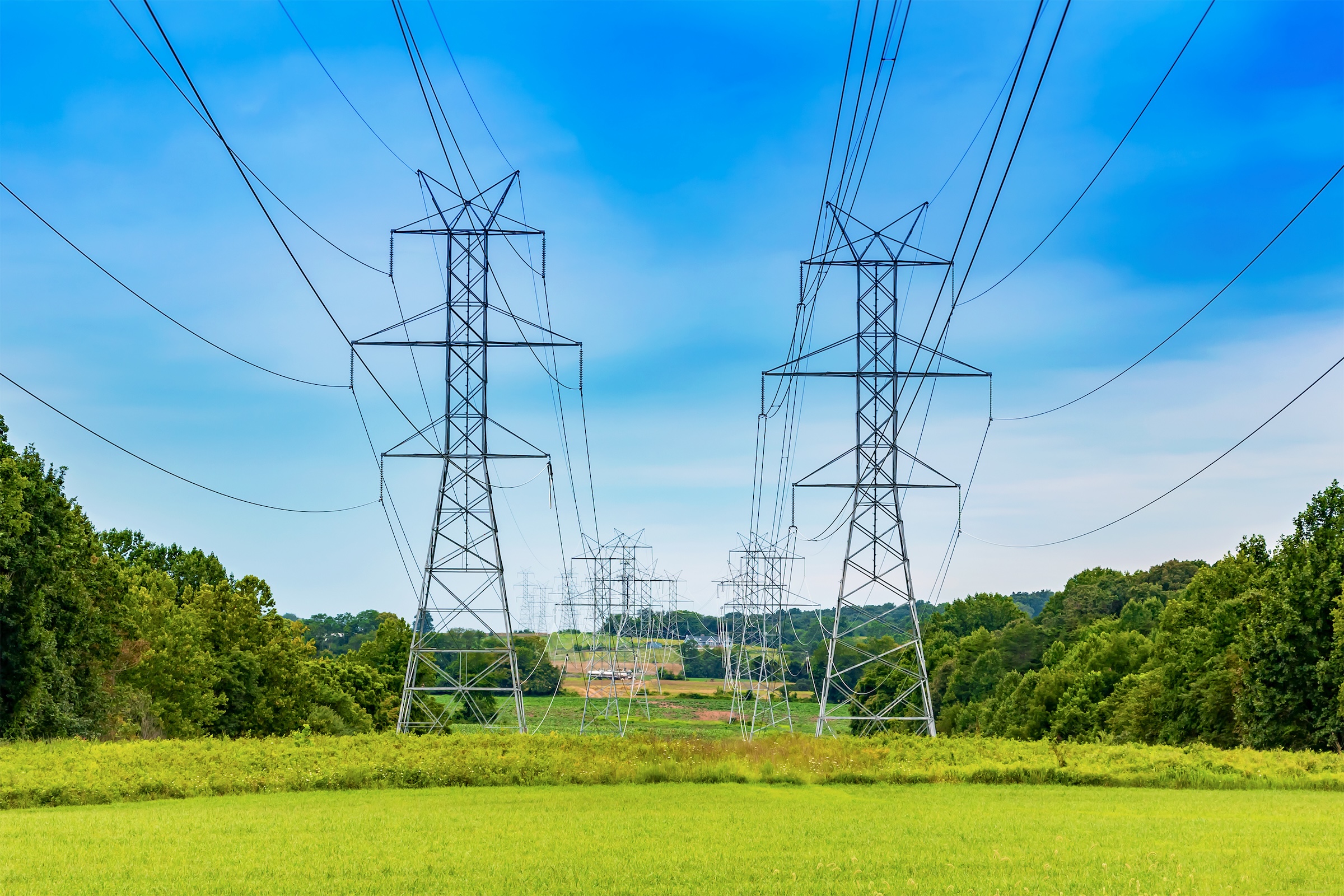 Steel electric transmission towers and overhead power lines extend across a green field bordered by trees under a blue sky, illustrating utility infrastructure passing through a rural landscape.
