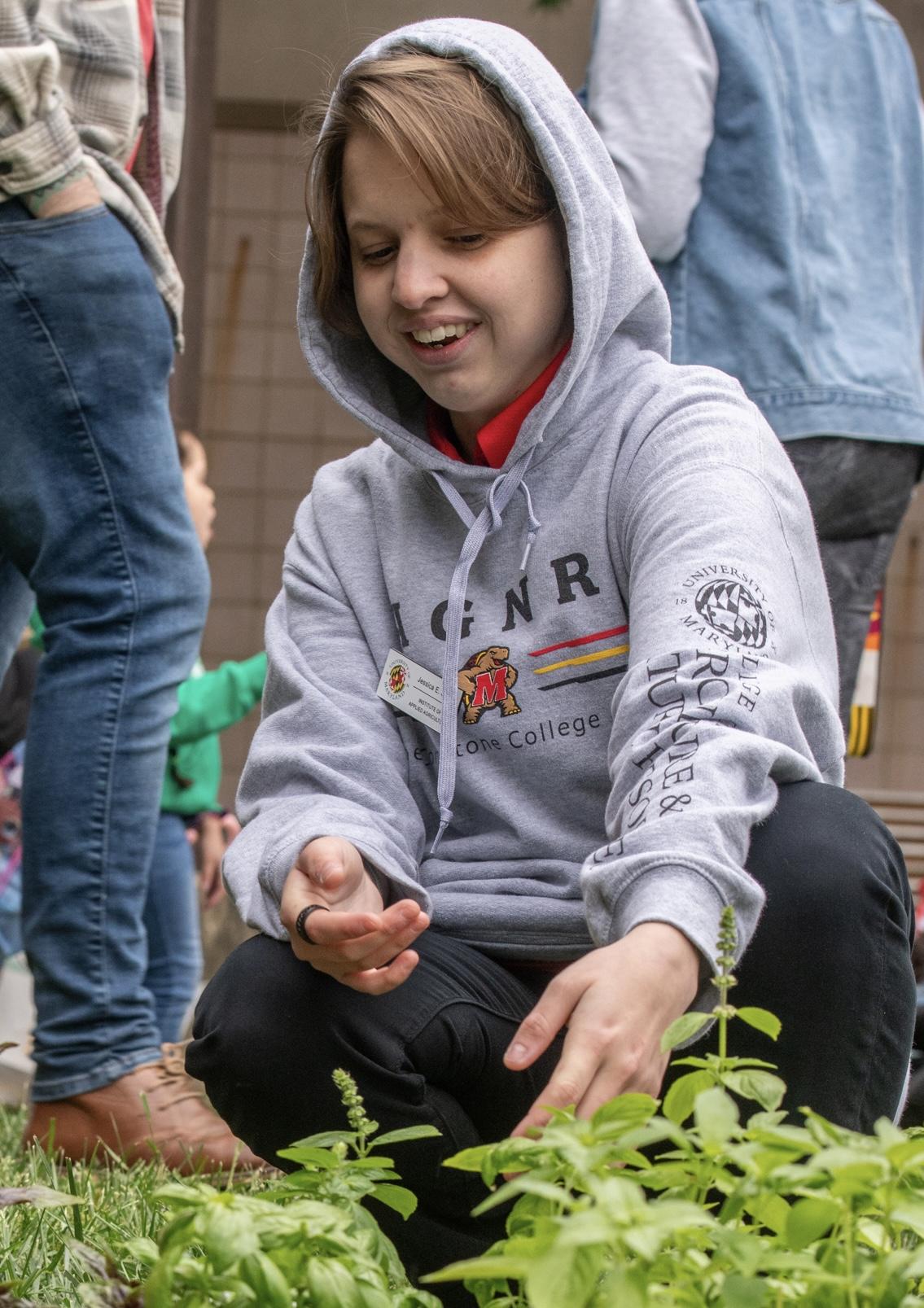 Student in a hooded sweatshirt kneeling to happily work with plants.