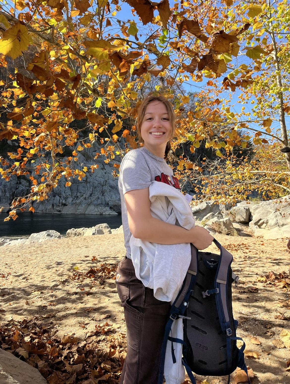 Happy student holding a backpack and surrounded by fall foliage. 