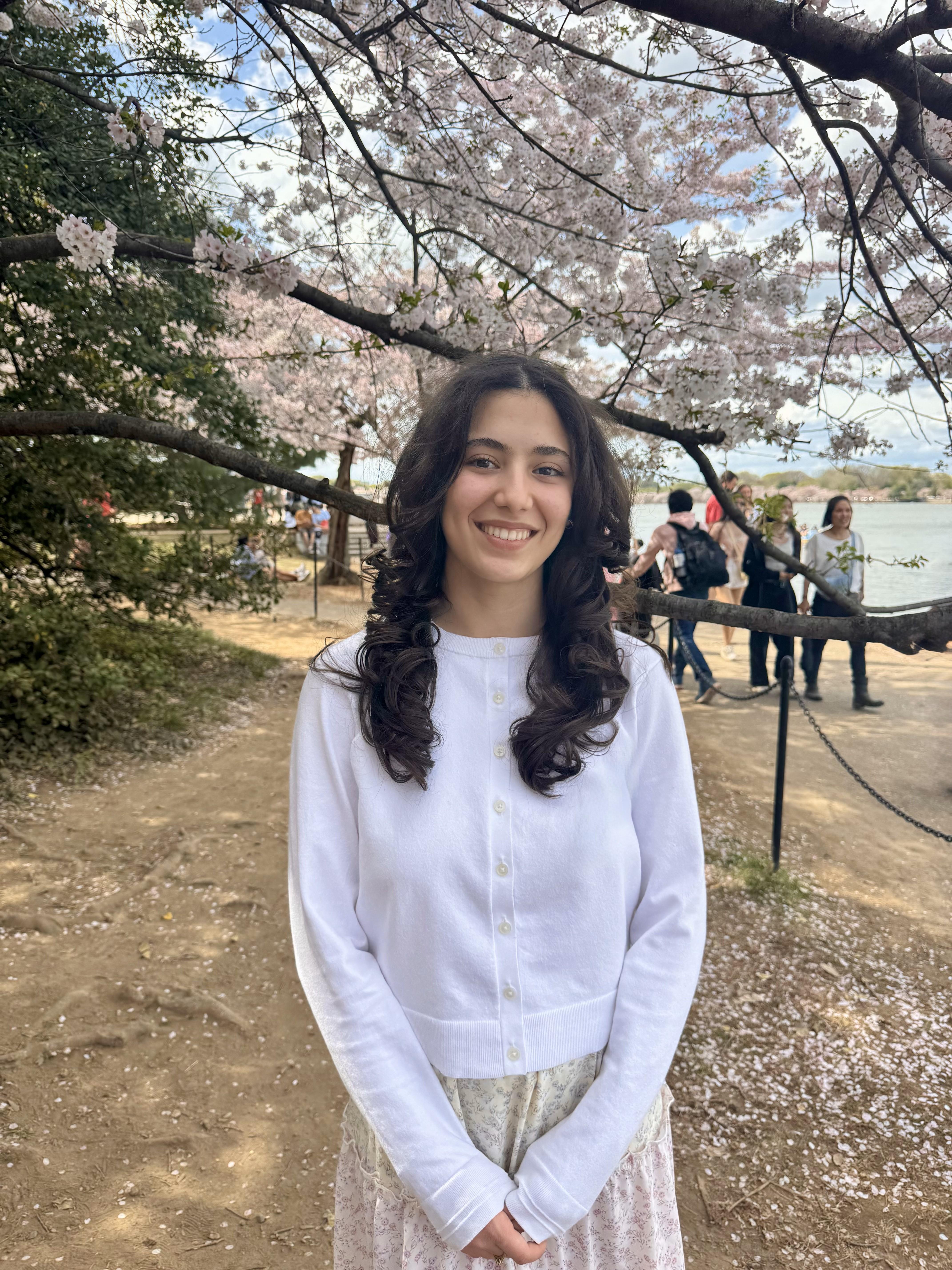 Student in a white cardigan standing in front of an evergreen and cherry blossom. 