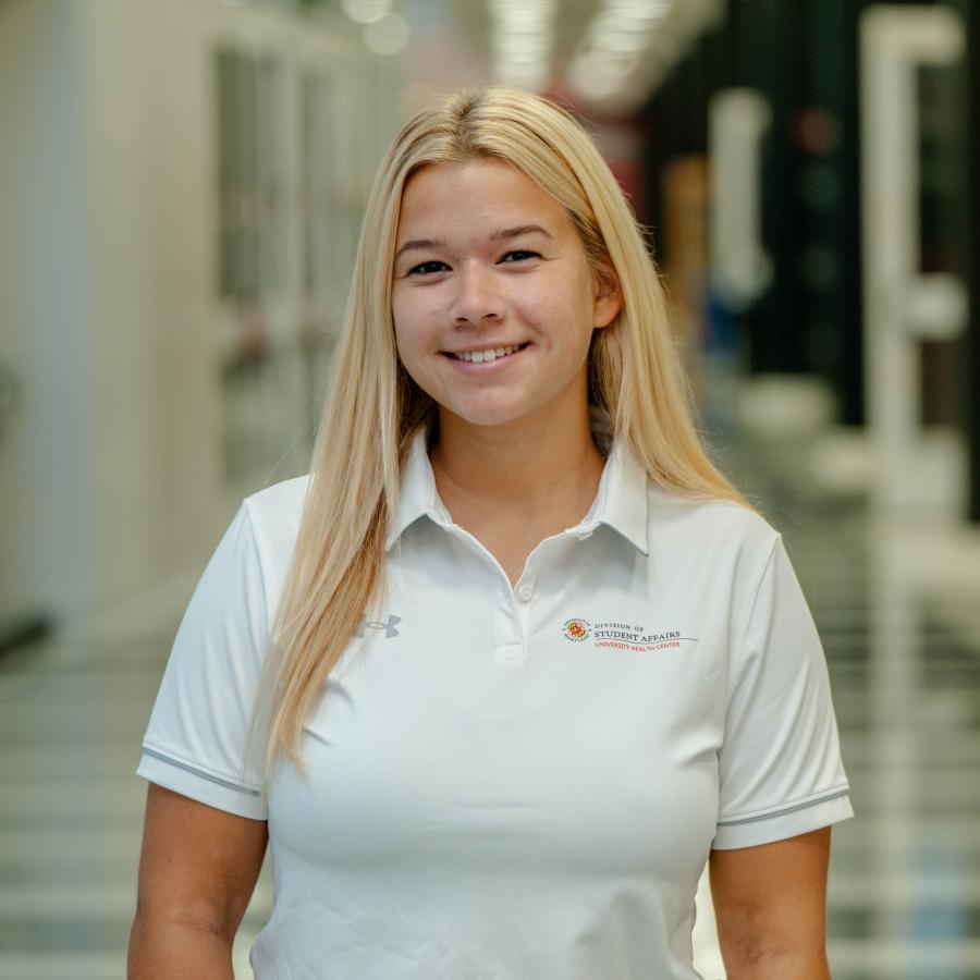 Cassidy is wearing a white polo and smiling for the camera with a long hallway in the background