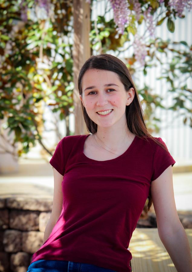 A smiling student dressed in a burgundy tshirt reclines against the backdrop of a young lilac.