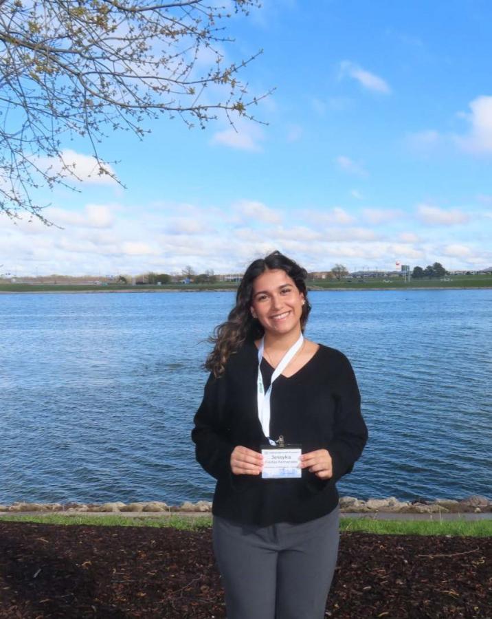 A student poses under a Cherry Blossom tree in front of the Potomac River, proudly displaying her professional conference nametag.