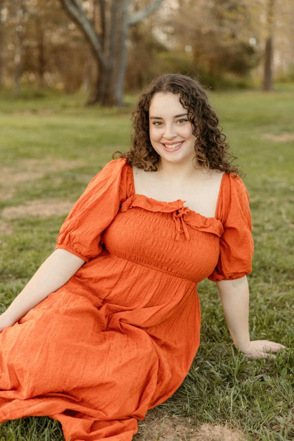 A student dressed in a vibrant tangerine colored dress reclining in a field of grass.