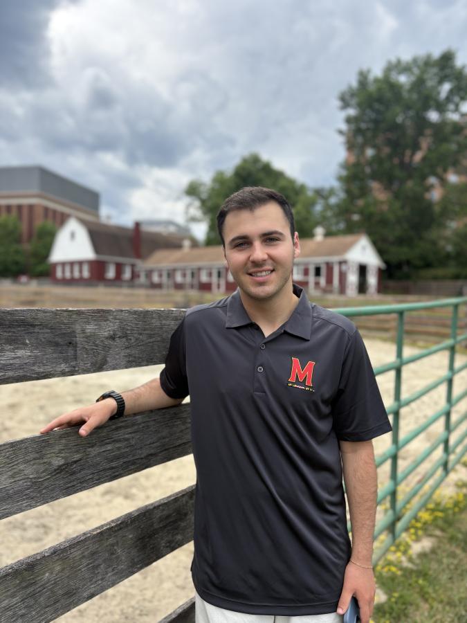 Smiling student standing against a fence at the on-campus University of Maryland farm proudly wears his UMD branded shirt.