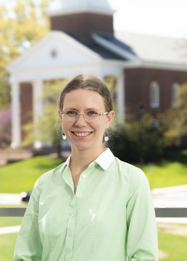 A student in a soft tea green shirt poses in front of an iconic University of Maryland building with the bright green lawn behind her accenting her smile.