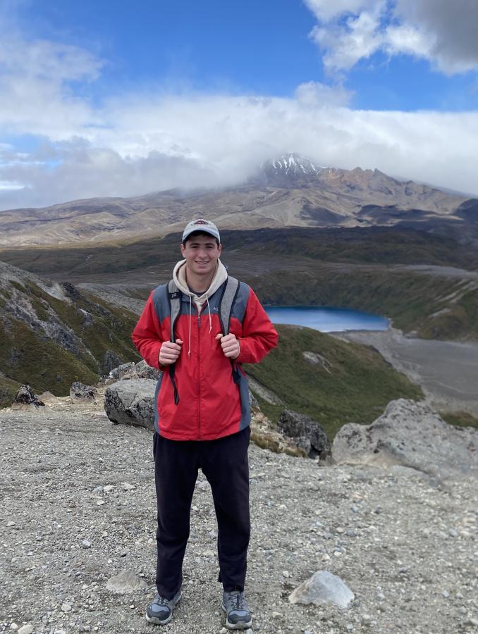 A student happily stands in the forefront of a sweeping vista with a bright blue sky mirrored in the glacial lake below.