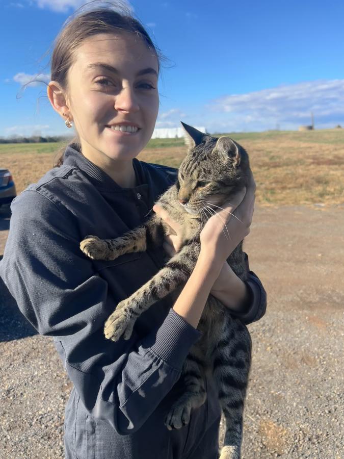A student in farm coveralls stands in the sunlight holding a grey tiger-striped cat.