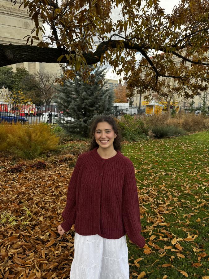 A smiling student wearing a long burgundy sweater standing amongst crisp fallen autumn foliage.