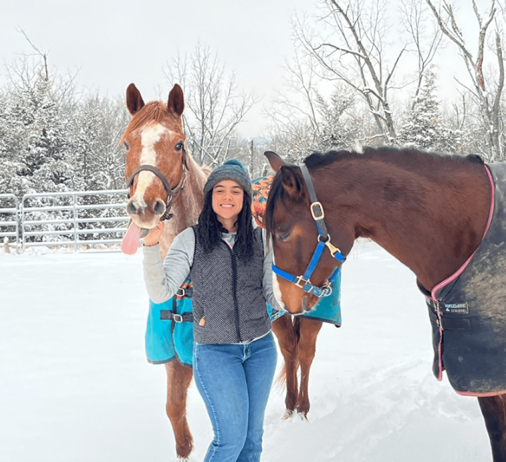 A student stands in a winter wonderland between two tall horses.